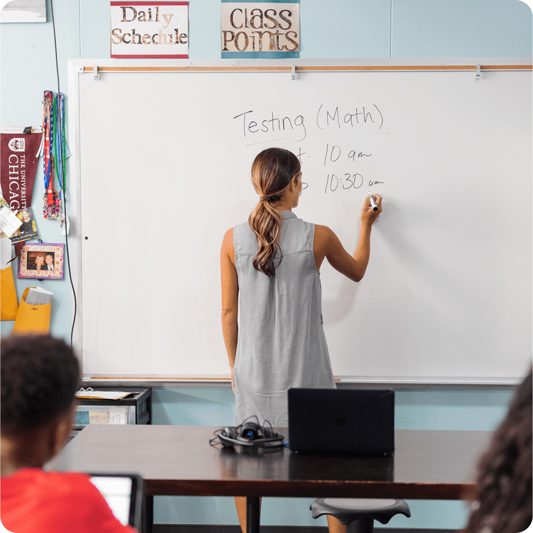 Teacher in classroom with whiteboard; desk featuring TWT Audio Duro headset and laptop.